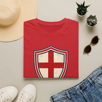 Thumbnail for Red t-shirt with a shield design on a beige surface with a hat, sunglasses, and shoes.