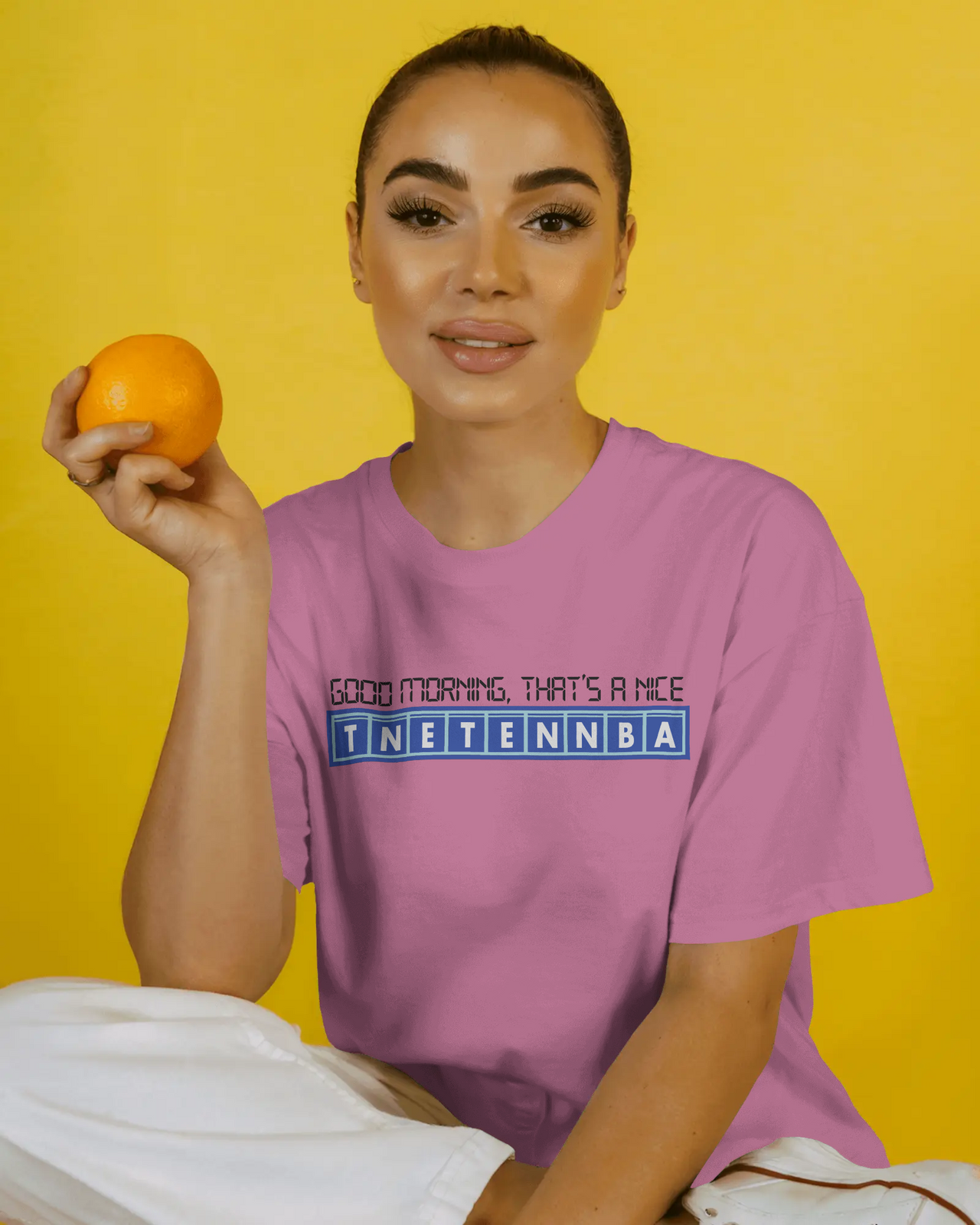 Woman holding an orange and wearing a pink t-shirt with text on a yellow background - Good Morning That’s A Nice TNETENNBA - The IT Crowd T-Shirt