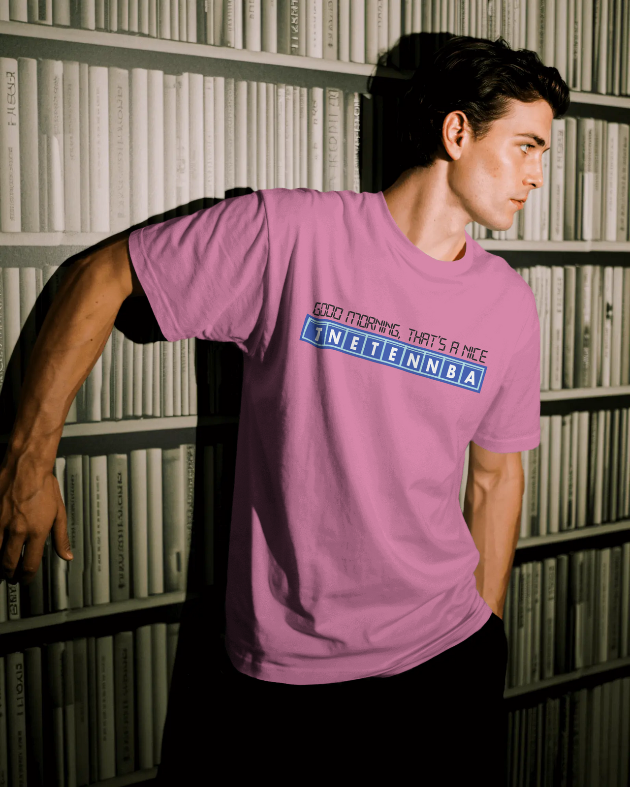 Person wearing a pink t-shirt with text in front of a bookshelf - Good Morning That’s A Nice TNETENNBA - The IT Crowd T-Shirt