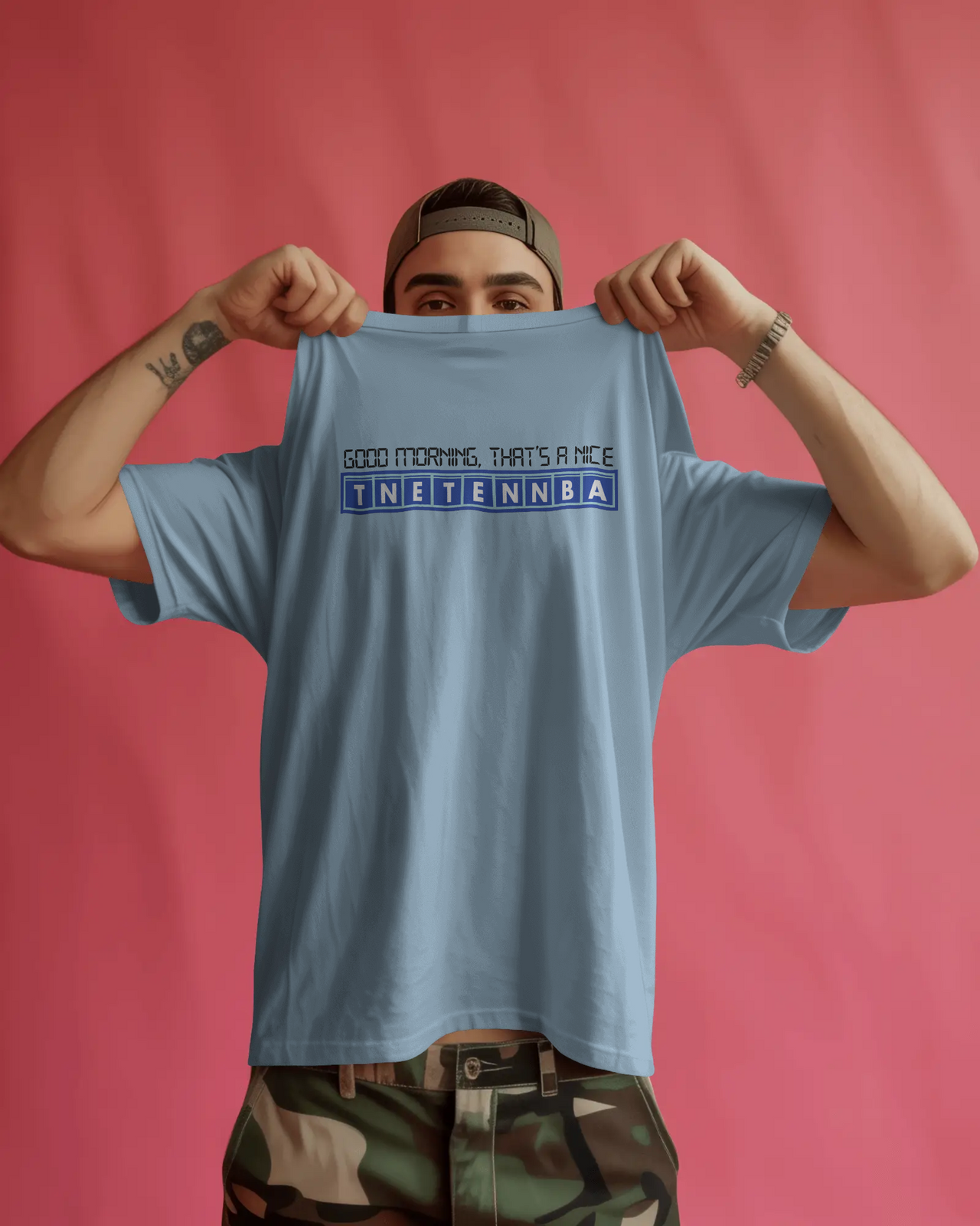Person holding up a light blue t-shirt with text against a pink background - Good Morning That’s A Nice TNETENNBA - The IT Crowd T-Shirt