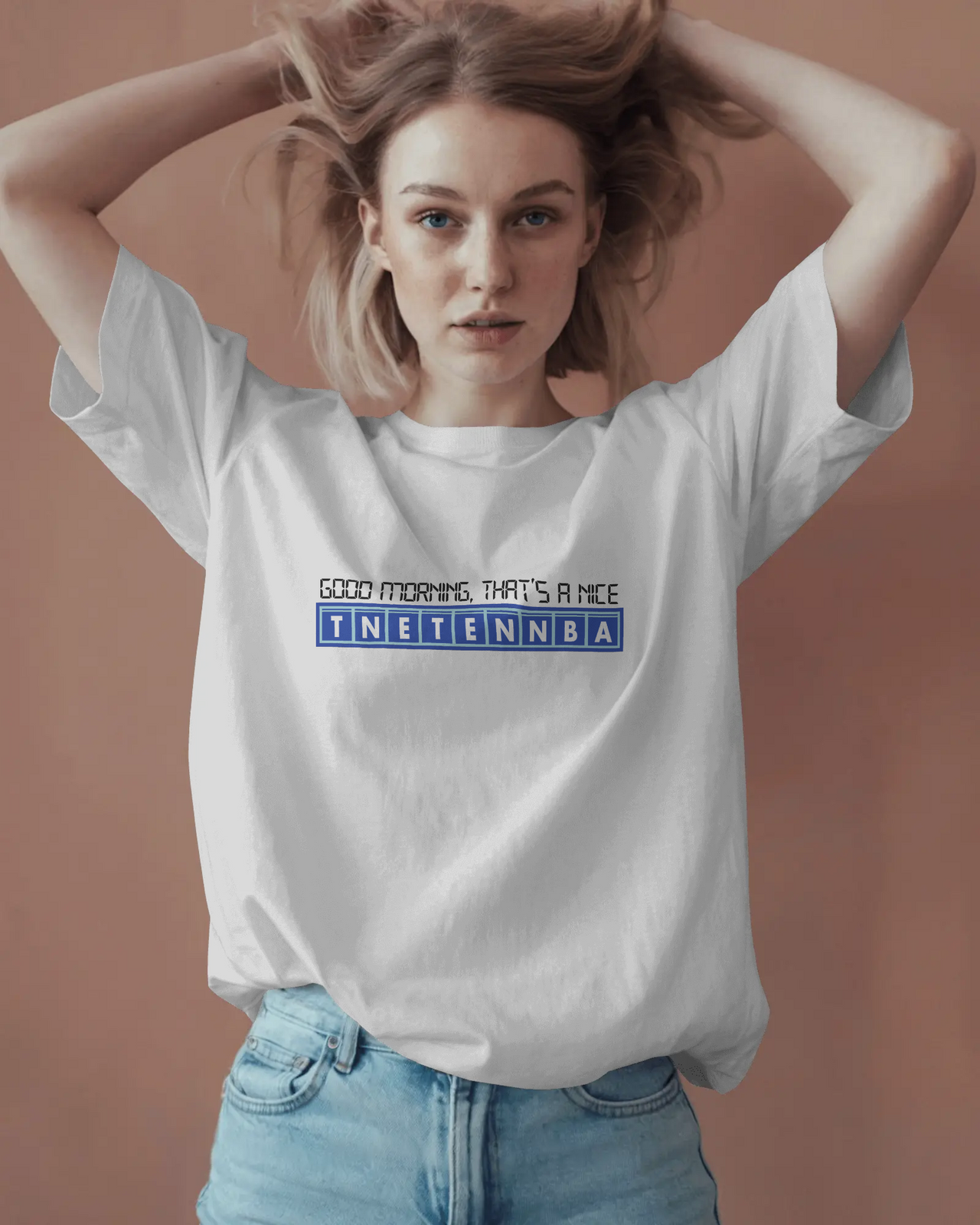 Woman wearing a white t-shirt with text on a beige background - Good Morning That’s A Nice TNETENNBA - The IT Crowd T-Shirt