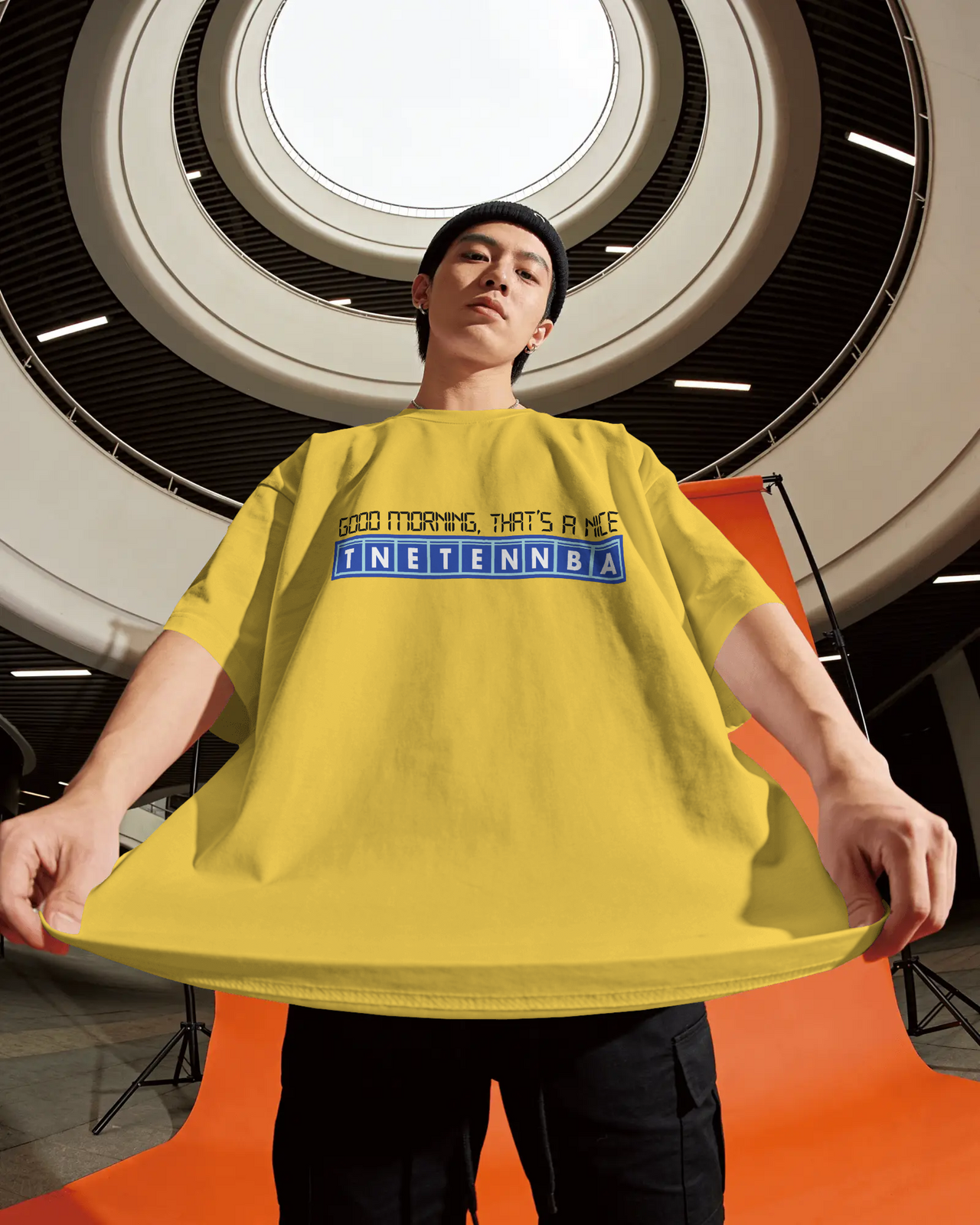 Person wearing a yellow oversized t-shirt with text in an indoor setting - Good Morning That’s A Nice TNETENNBA - The IT Crowd T-Shirt