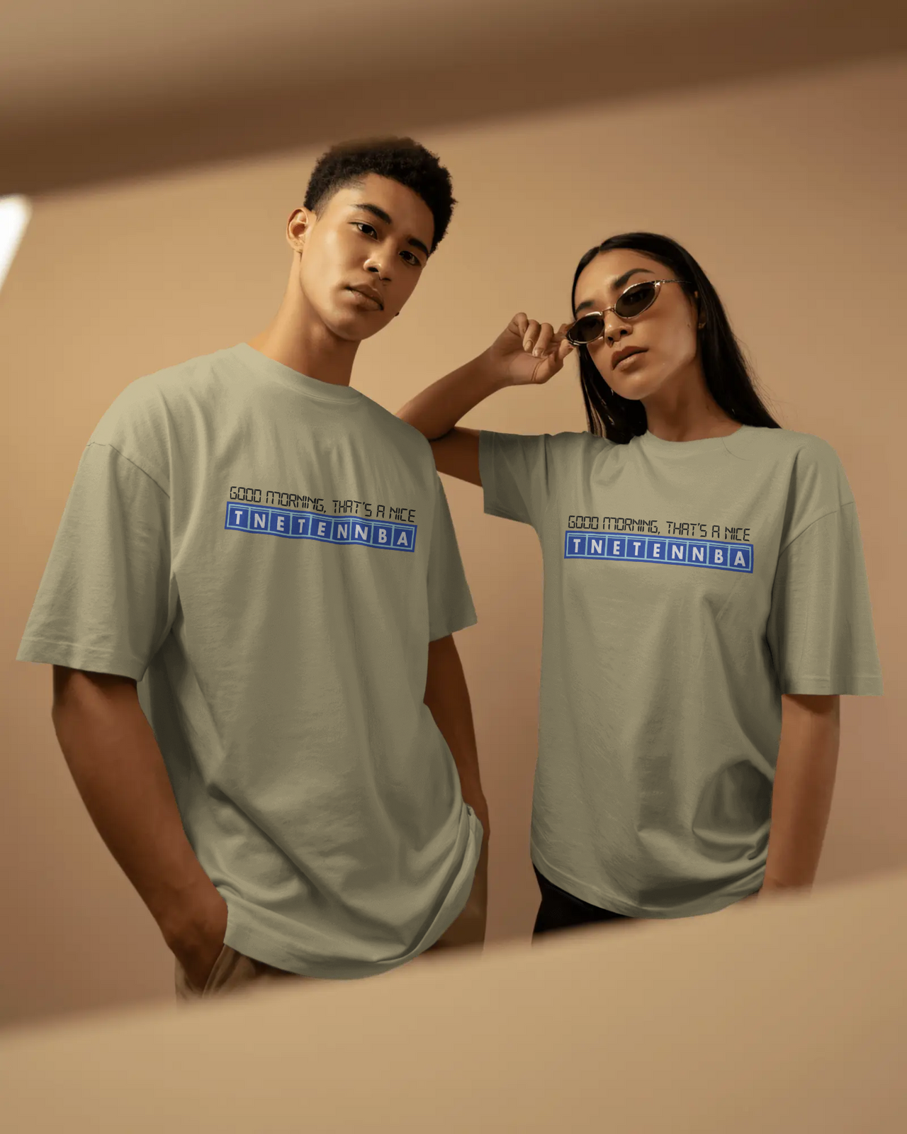 Two people wearing matching light gray t-shirts with a blue and white logo on a beige background - Good Morning That’s A Nice TNETENNBA - The IT Crowd T-Shirt