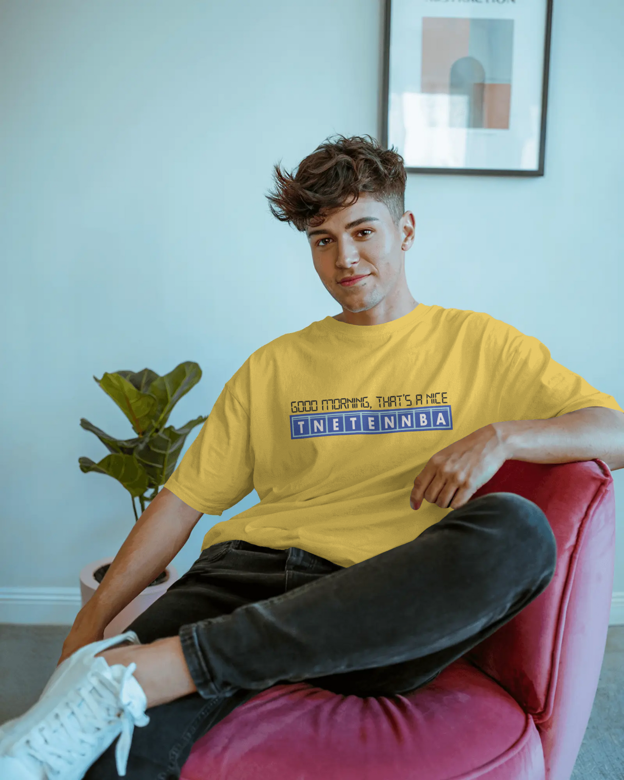 Person wearing a yellow t-shirt with text, sitting on a red chair in a room with a plant and framed picture. - IT Crowd - Tnetennba