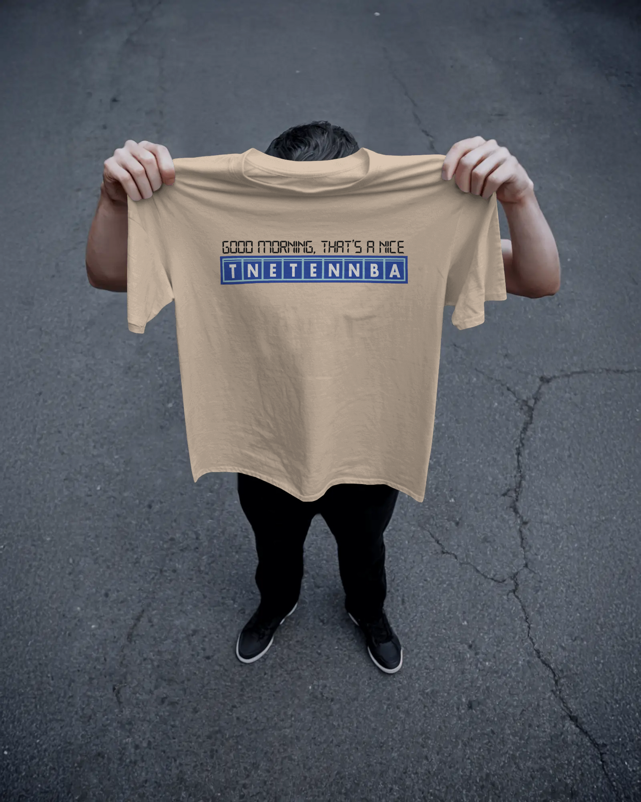Person holding a beige t-shirt with text on a dark asphalt background - Good Morning That’s A Nice TNETENNBA - The IT Crowd T-Shirt