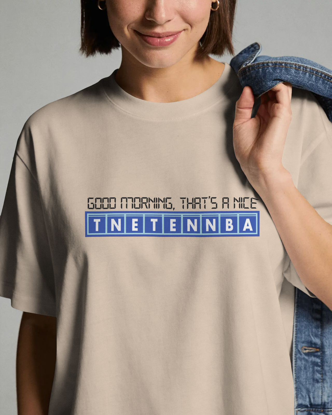 Person wearing a beige t-shirt with text and a logo, holding jeans. - IT Crowd - Tnetennba
