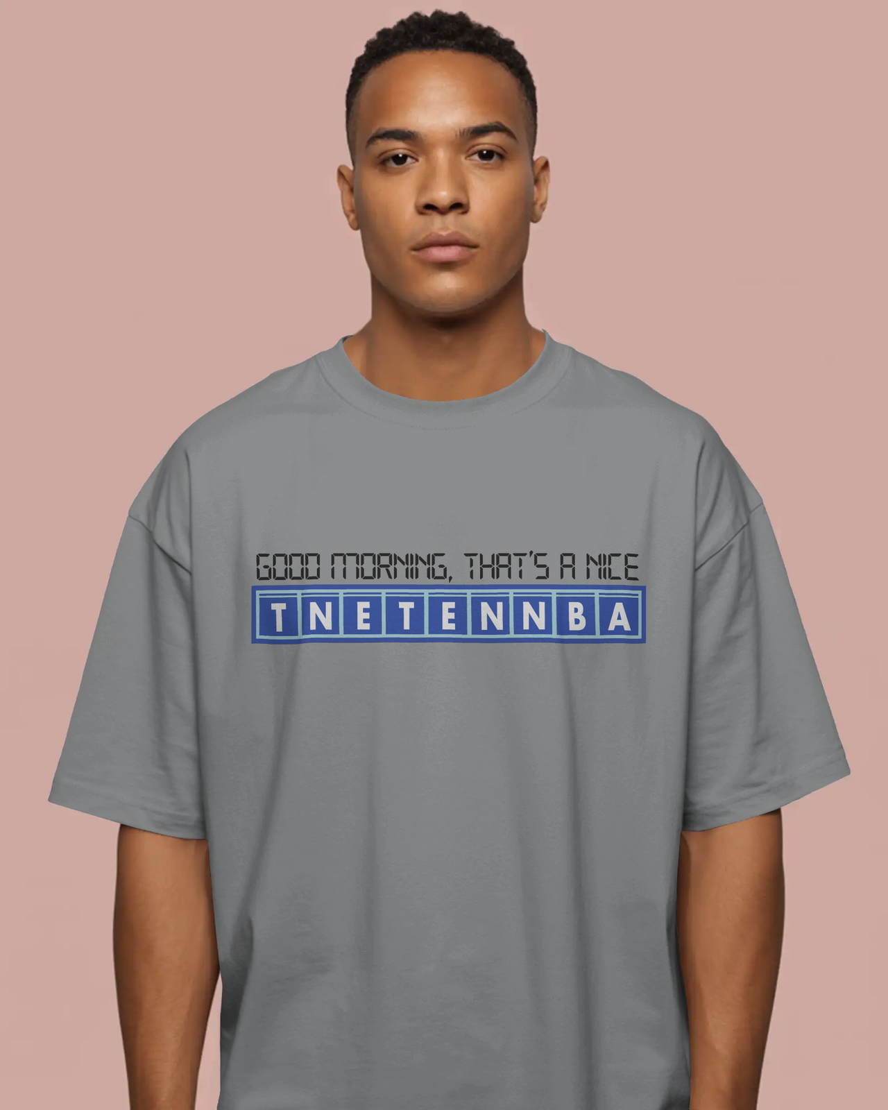 Man wearing a gray t-shirt with text on a pink background - Good Morning That’s A Nice TNETENNBA - The IT Crowd T-Shirt