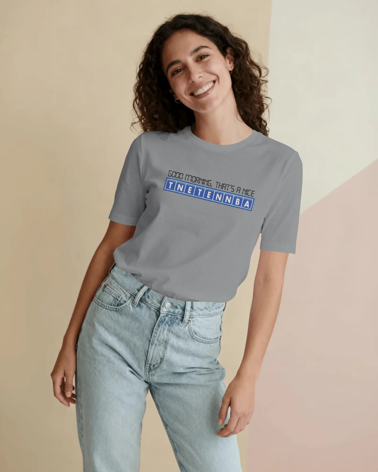 Person wearing a gray t-shirt with text and light blue jeans against a beige background - Good Morning That’s A Nice TNETENNBA - The IT Crowd T-Shirt