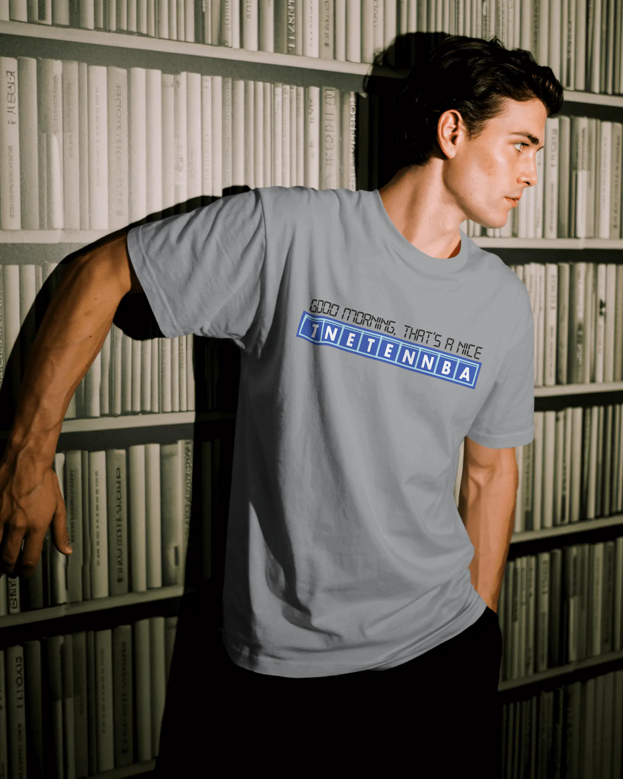 Person wearing a gray t-shirt with text on the back in front of a bookshelf. - Good Morning That’s A Nice TNETENNBA - The IT Crowd T-Shirt