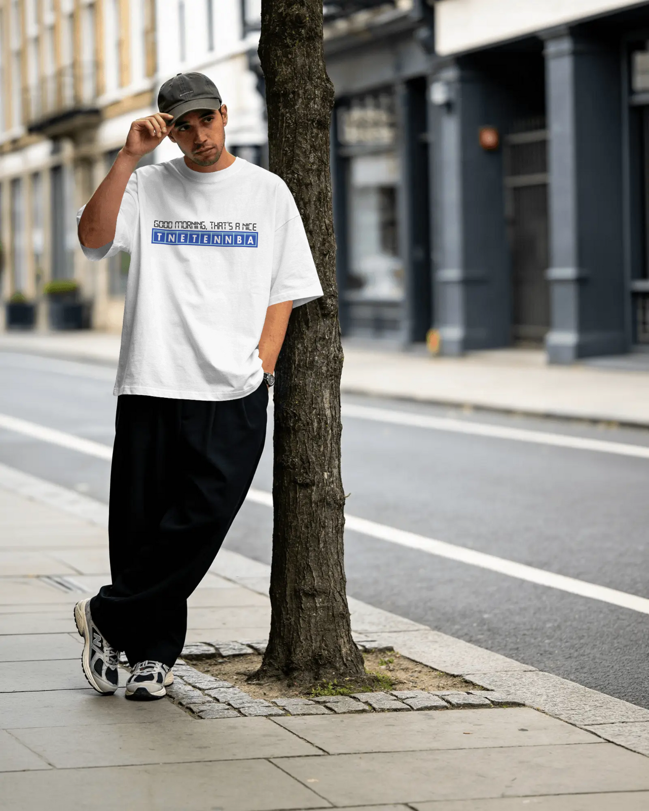 Person wearing a white t-shirt with text, black pants, and a cap, standing on a city street. - IT Crowd - Tnetennba