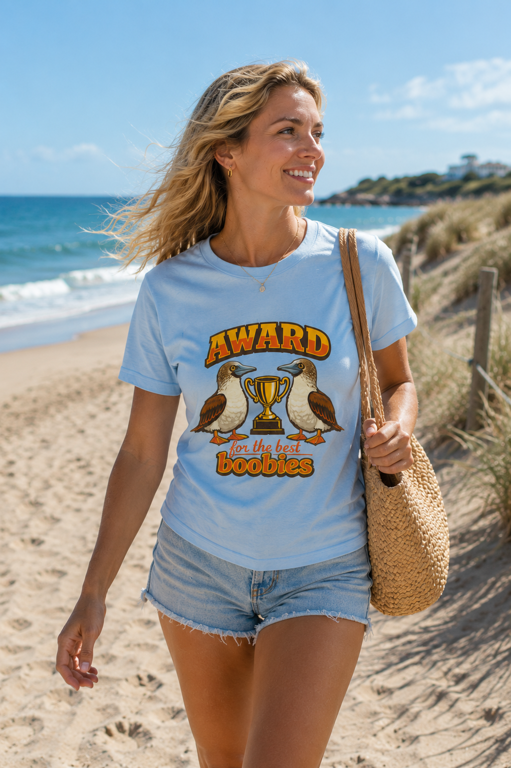 Woman wearing the Award for the Best Boobies T-Shirt in Light Blue while walking beside beach dunes on a sunny day.
