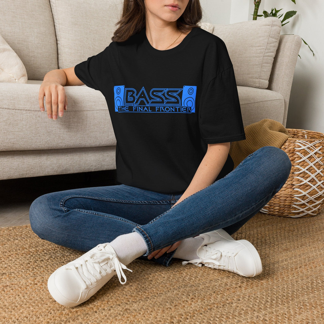 A woman sitting cross-legged on a woven rug in front of a beige sofa, wearing a black graphic T-shirt with a blue 'BASS THE FINAL FRONTIER' design, blue jeans, and white sneakers.