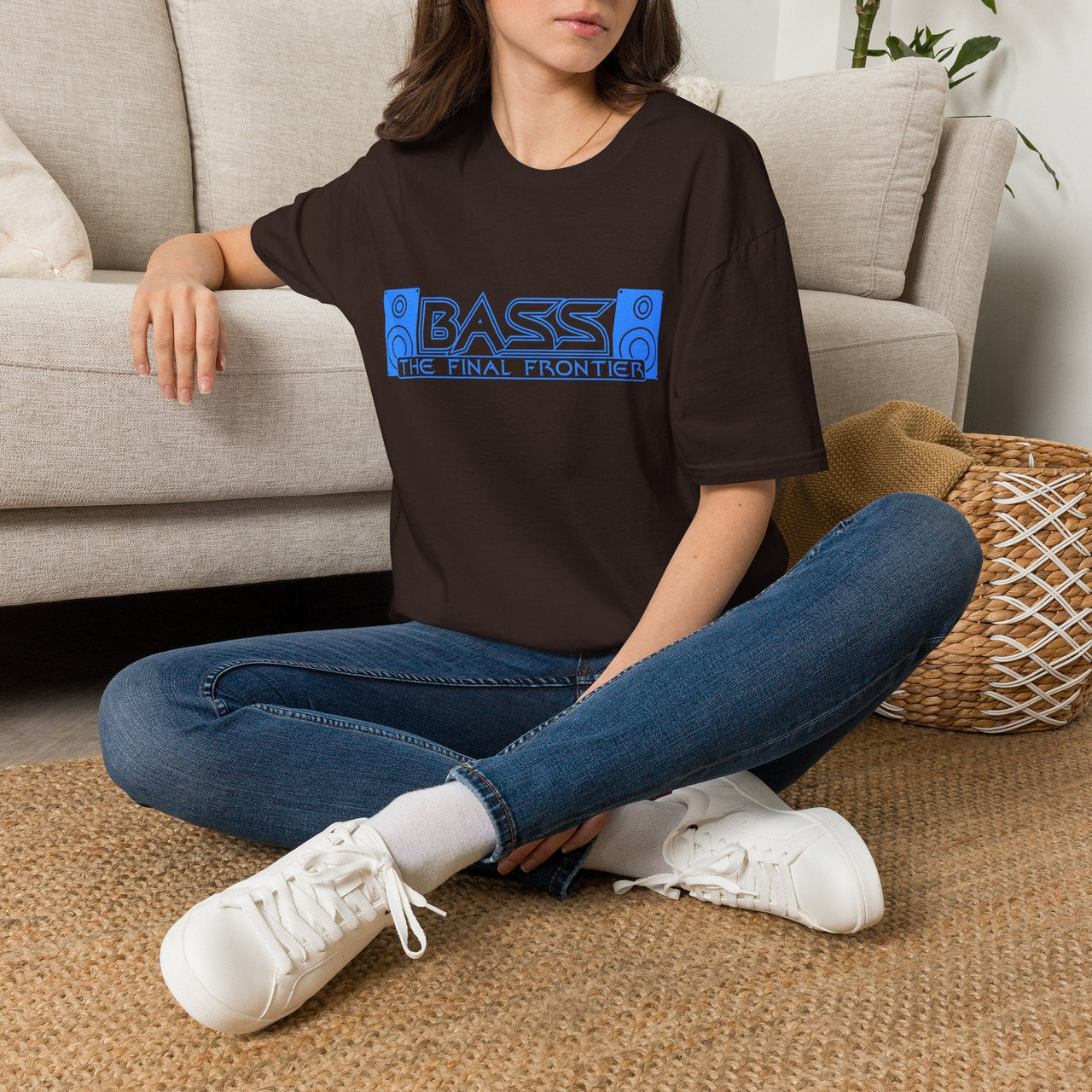 A woman sitting cross-legged on a woven rug in front of a beige sofa, wearing an oversized dark brown t-shirt with a blue graphic that reads 'BASS - THE FINAL FRONTIER' and blue jeans with white sneakers.