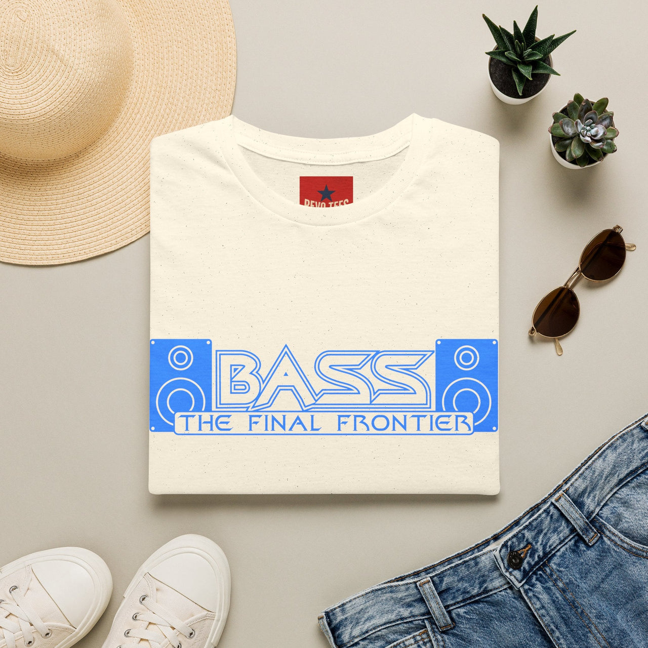A folded natural-colored t-shirt with a blue graphic reading 'BASS THE FINAL FRONTIER' is styled in a flat lay with a straw hat, white sneakers, blue jeans, sunglasses, and small potted succulents on a light gray background.