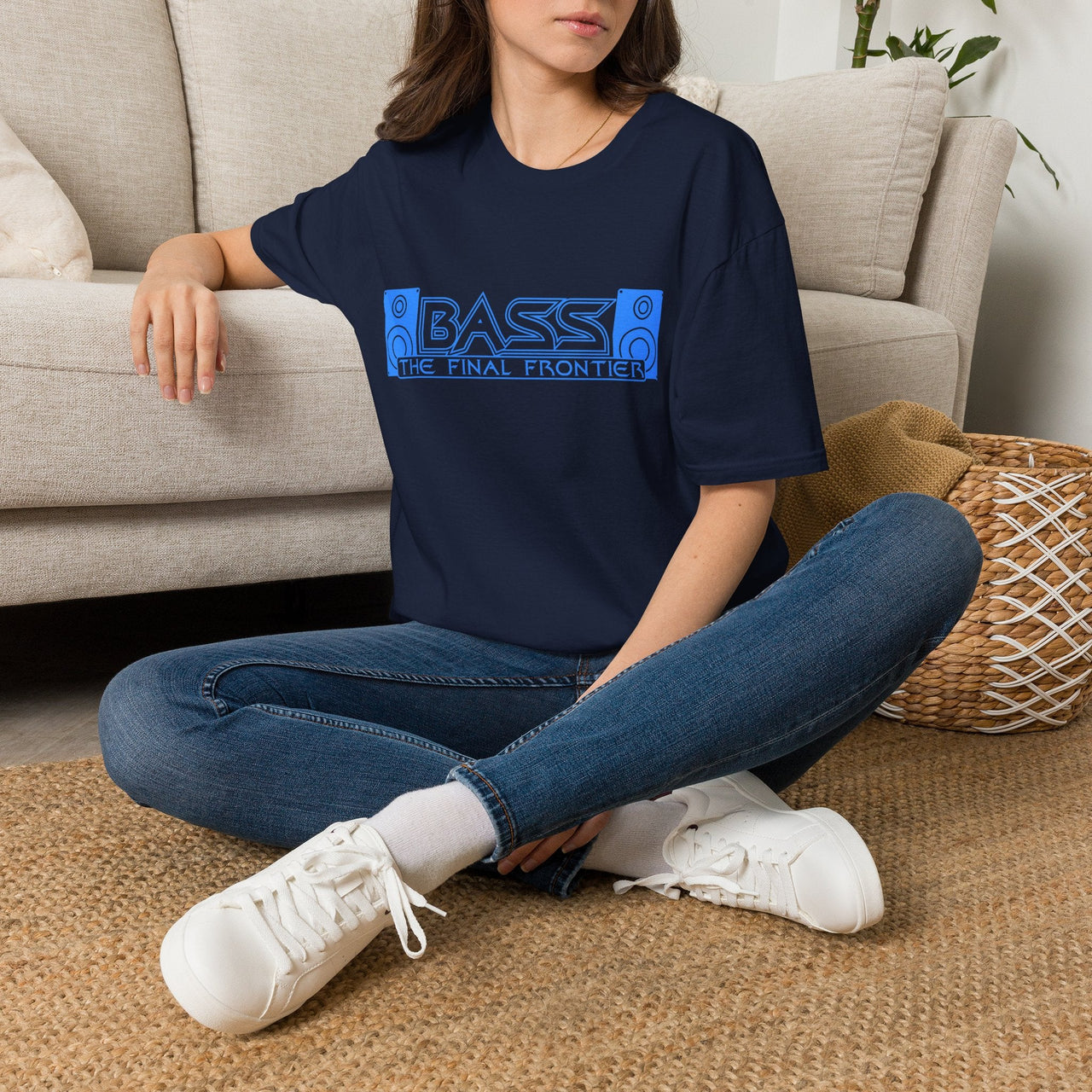 A woman sitting cross-legged on a woven rug in front of a beige sofa, wearing a navy blue graphic T-shirt with a blue 'BASS THE FINAL FRONTIER' design, blue jeans, and white sneakers.