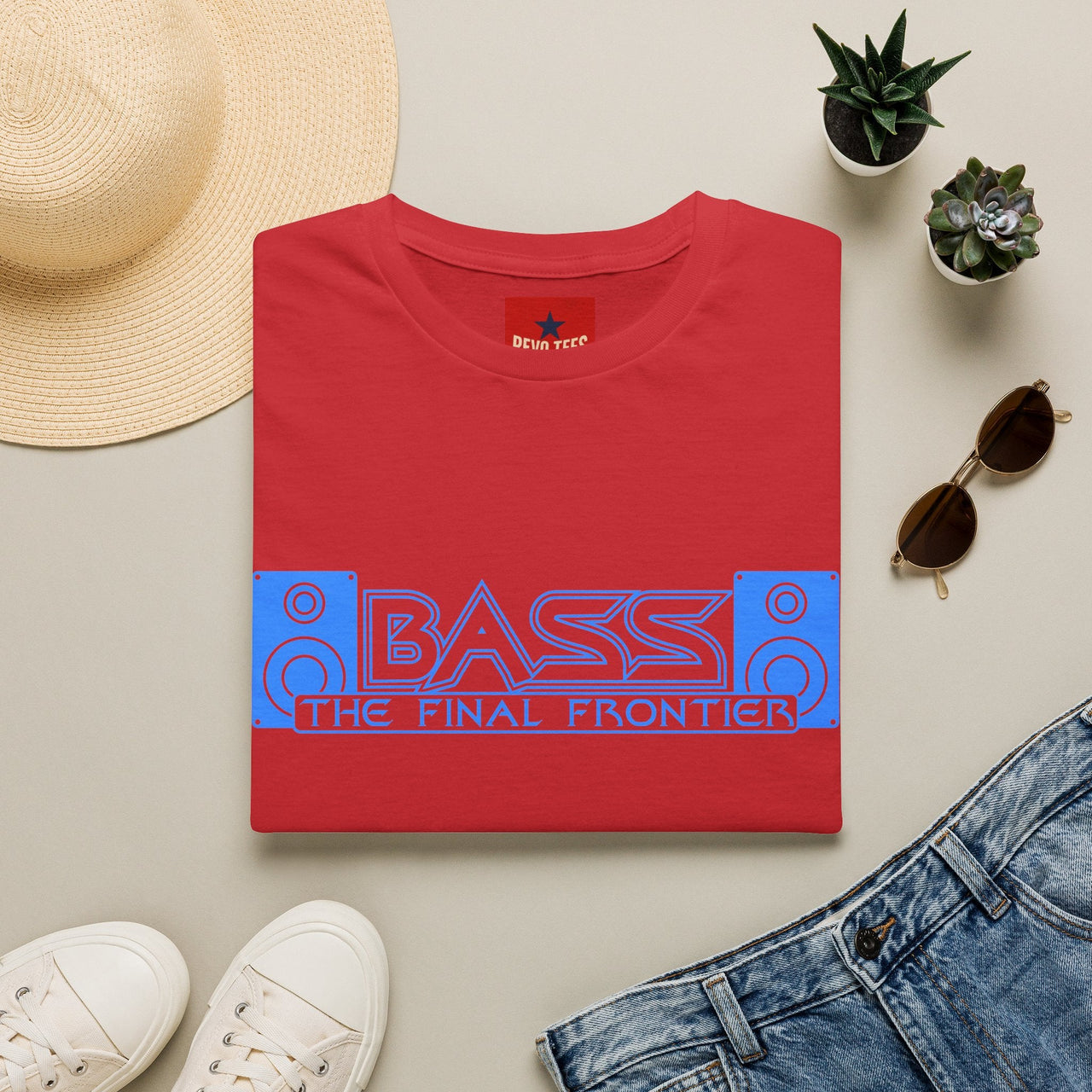 A folded red graphic T-shirt is arranged in a styled flat lay on a light beige surface, surrounded by a straw hat, white sneakers, blue jeans, small potted succulents, and sunglasses. The shirt has a large blue and red retro speaker graphic with the text 'BASS THE FINAL FRONTIER' across the chest.