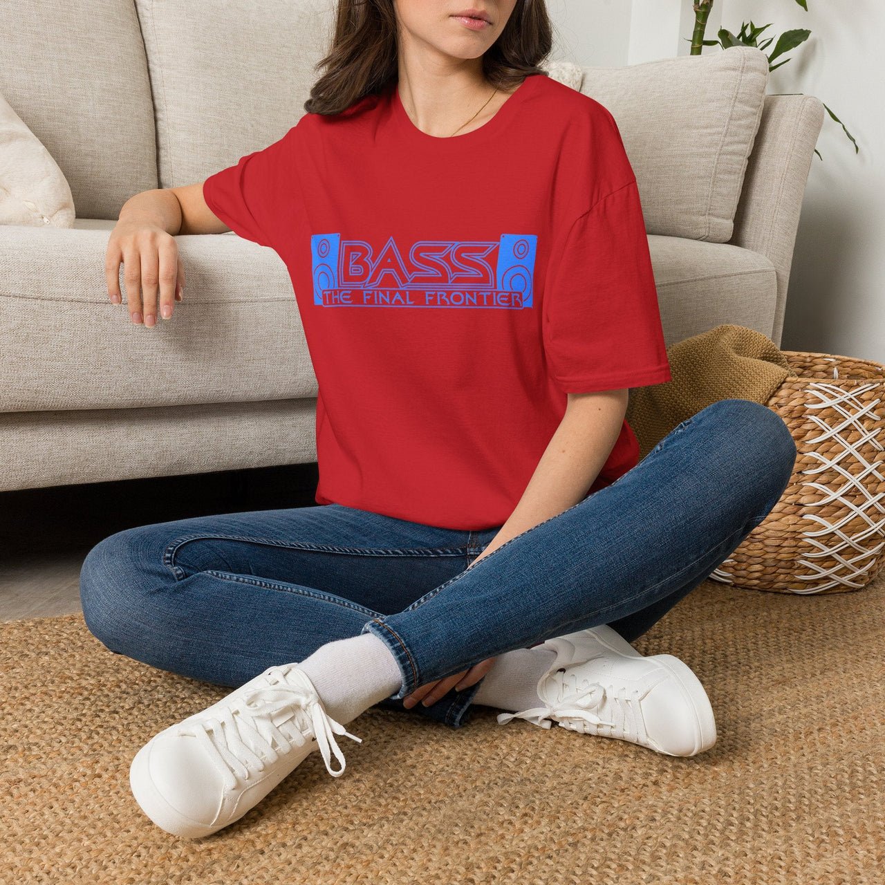 A woman sitting cross-legged on a woven rug in front of a light beige sofa, wearing a red graphic t-shirt with blue and pink text that reads 'BASS - THE FINAL FRONTIER', blue jeans, white sneakers, and white socks.