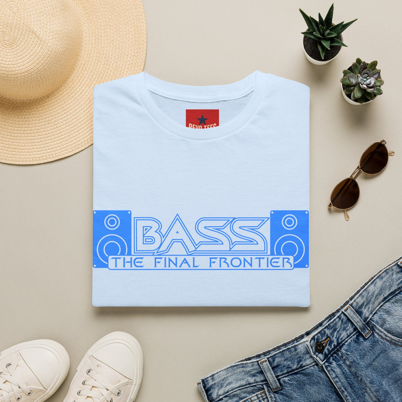 White t-shirt with 'BASS THE FINAL FRONTIER' design, placed on a beige surface with a hat, sunglasses, and jeans.