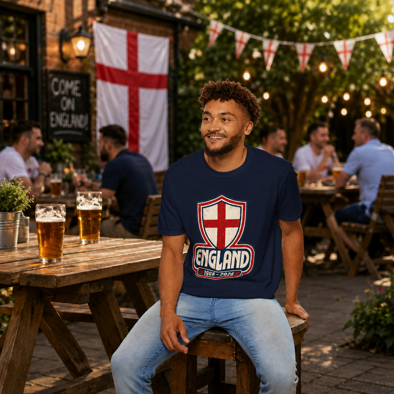 Man wearing the England 1966–2026 Retro St George Cross T-Shirt in a pub garden with England flags in the background.
