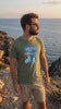 Man wearing a Military Green Nuke The Whales T-Shirt by the coast in a short product video, with the retro cartoon-style graphic clearly visible.