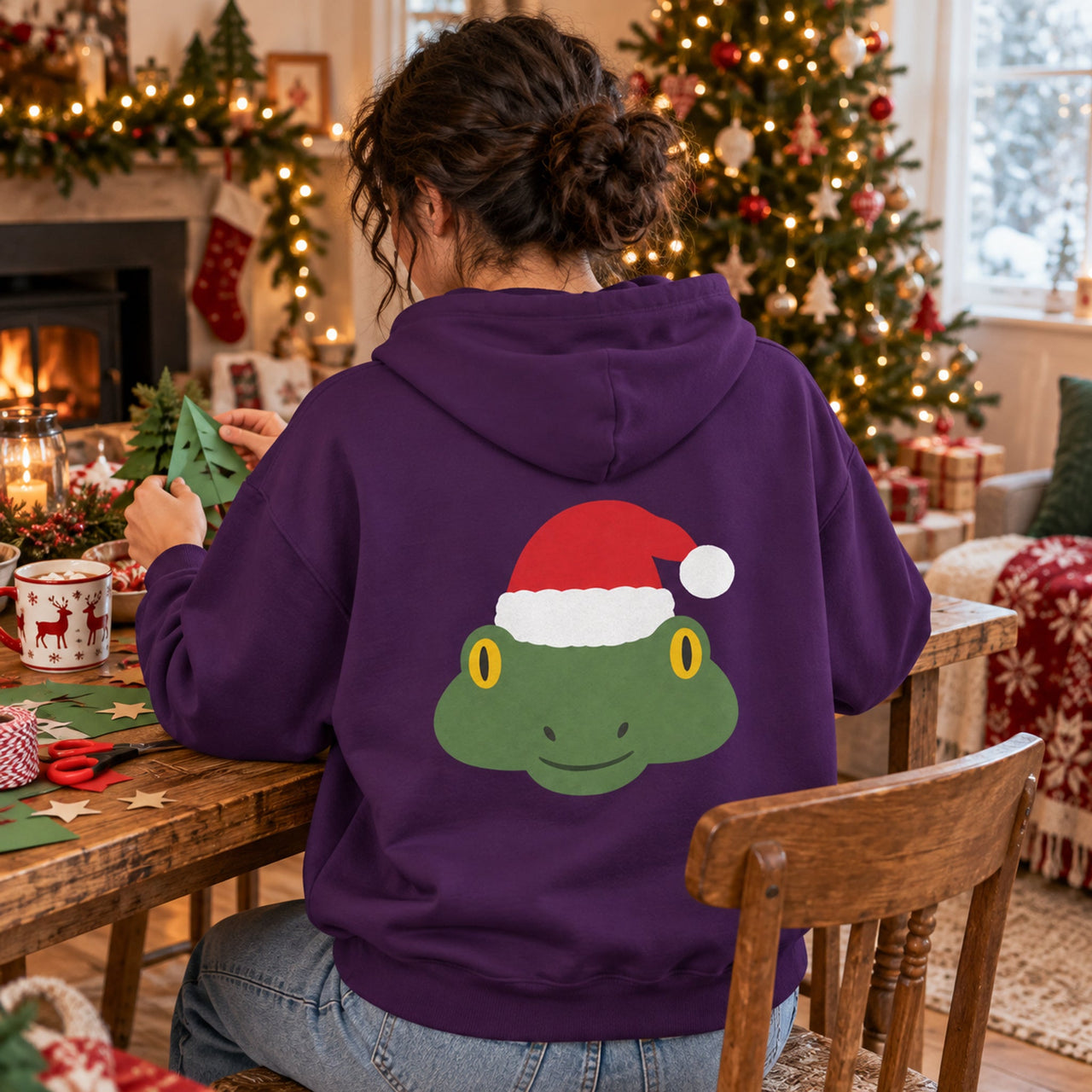 Santa Frog Christmas Back Print Hoodie in Purple, worn in a Christmas home setting with the Santa frog back print visible.