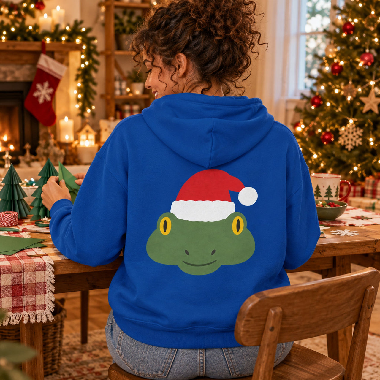 Santa Frog Christmas Back Print Hoodie in Royal, worn in a Christmas home setting with the Santa frog back print visible.