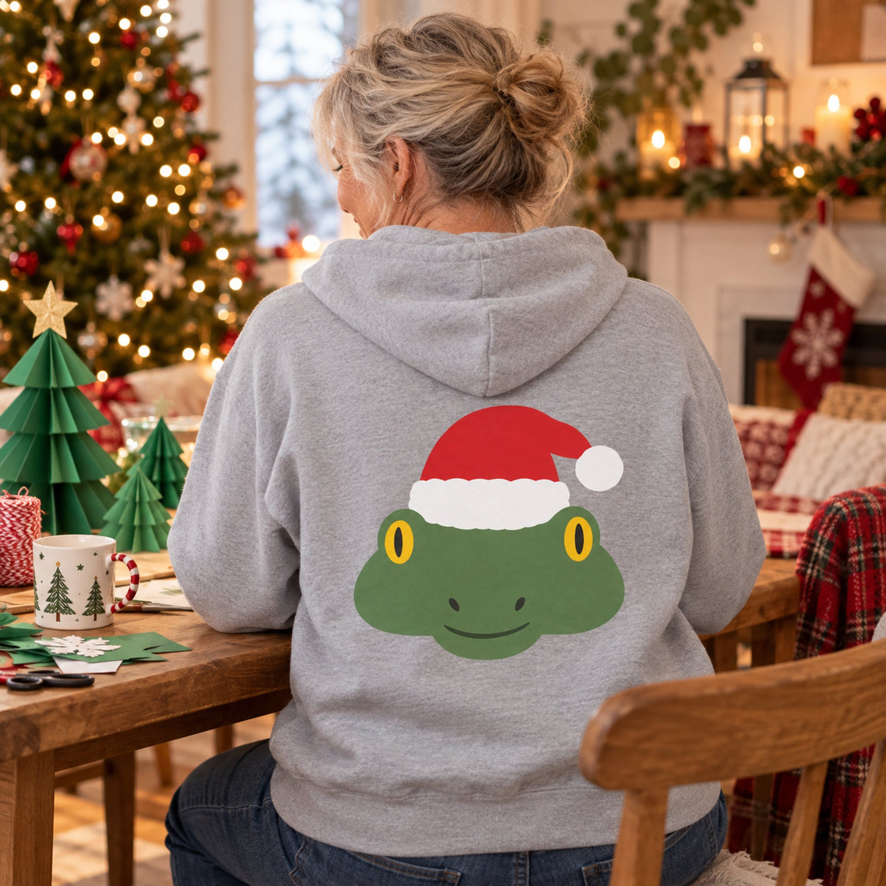 Santa Frog Christmas Back Print Hoodie in Sport Grey, worn in a Christmas home setting with the Santa frog back print visible.